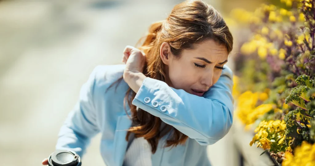 Una mujer con una chaqueta azul claro está estornudando y cubriéndose la nariz con el brazo, con flores amarillas en el fondo que sugieren alergias estacionales.