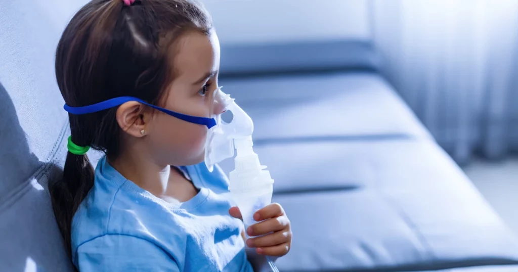 Una niña con el cabello oscuro recogido hacia atrás usa una máscara nebulizadora mientras está sentada en un sofá, mirando hacia un lado, con una suave iluminación interior.
