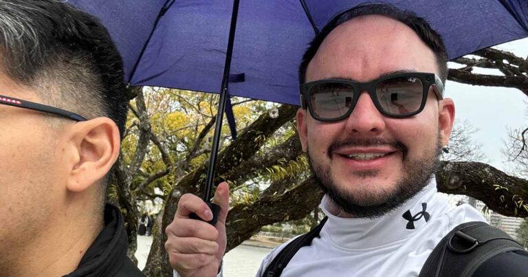 Un hombre con gafas de sol y una chaqueta blanca de Under Armour sonríe a la cámara mientras sostiene un paraguas azul. Otra persona está de pie junto a él, parcialmente fuera de plano. Al fondo se ven árboles sin hojas.