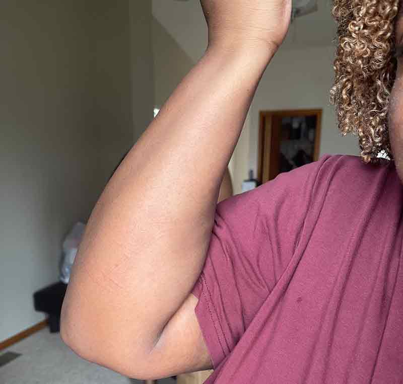 Una persona que viste una camisa marrón, con cabello rizado visible, levanta su brazo izquierdo, mostrando la parte interna del antebrazo suave. El fondo incluye una pared beige, una puerta y algunos artículos del hogar.