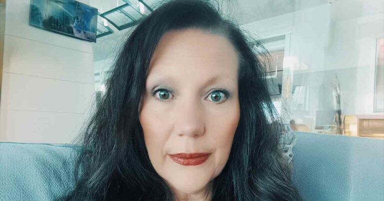 Una mujer con cabello largo y oscuro y ojos claros está sentada en un sofá azul claro en el interior, mirando directamente a la cámara con una expresión neutra.