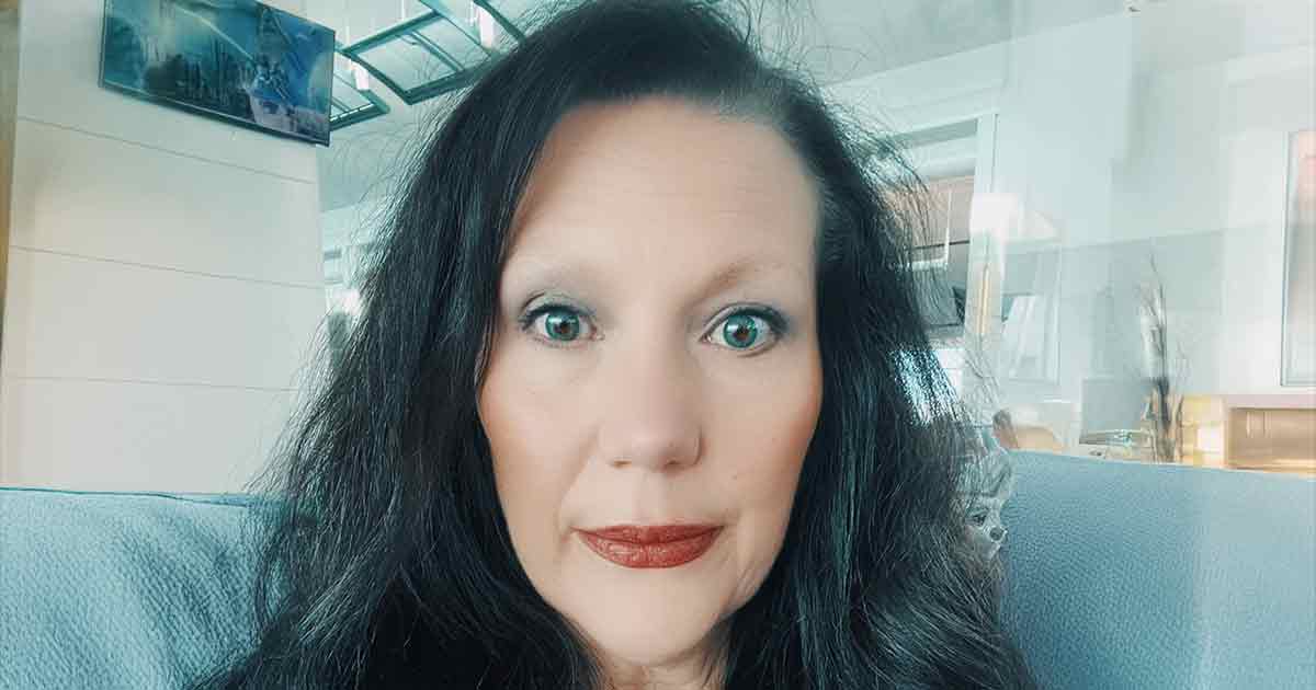 Una mujer con cabello largo y oscuro y ojos claros está sentada en un sofá azul claro en el interior, mirando directamente a la cámara con una expresión neutra.